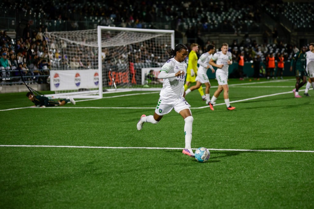 Greenville Triumph player in the white away kit carrying the ball against AV ALTA FC at Lancaster Municipal Stadium